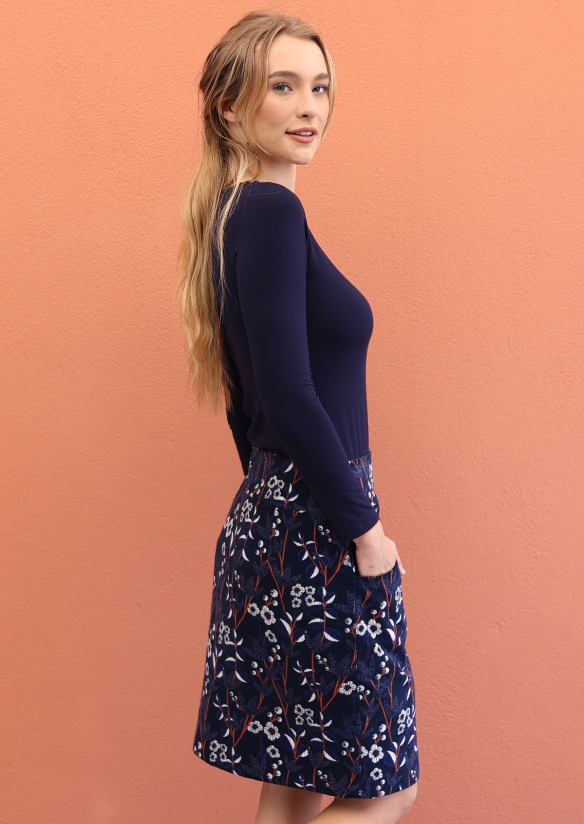 model wearing a mid length skirt featuring a meadow flower print with blue, white and orange on a navy blue base.