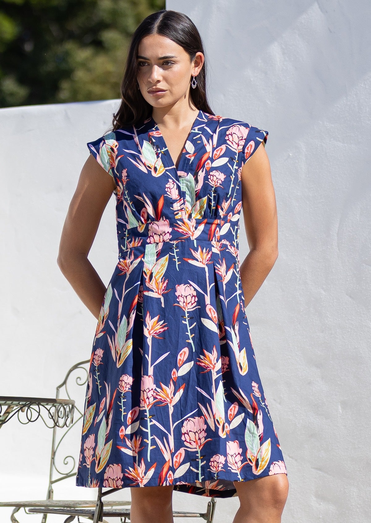 Woman wears above knee length cotton dress with V neckline, cap sleeves and softly cinched waist.