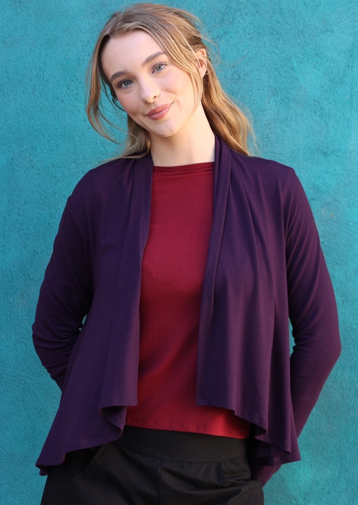 Model wears dark purple long sleeve stretch rayon cardi with waterfall front hem that exposes maroon top and black pants underneath