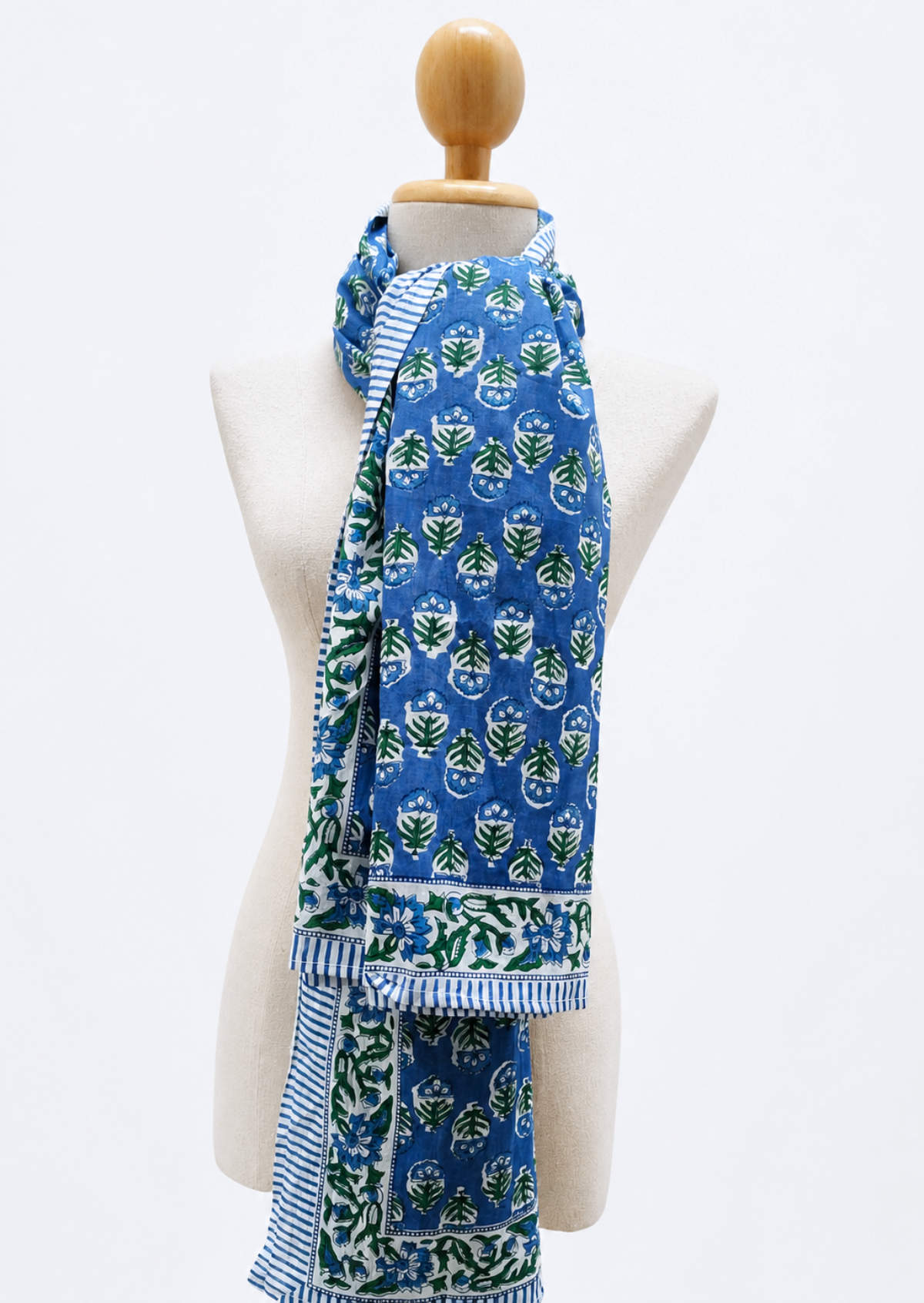 A blue scarf with green and white block-printed floral motifs and striped borders, draped on a cream-colored mannequin torso against a white background.