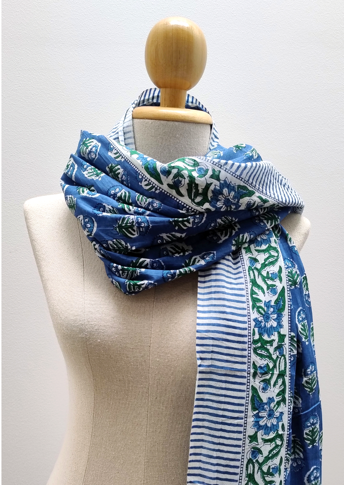 A blue patterned scarf with green floral motifs and white stripes, draped on a cream-colored fabric mannequin torso with a wooden neck cap.
