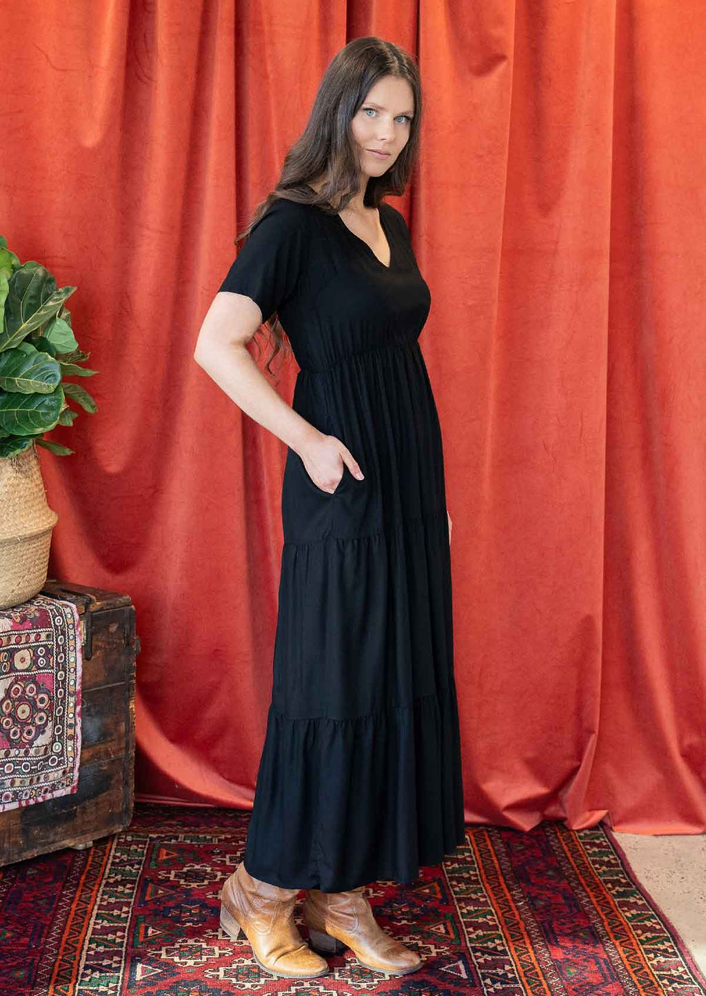 Woman standing side on in a black rayon maxi dress with short sleeves and a V-neckline, with her hand in side poceket