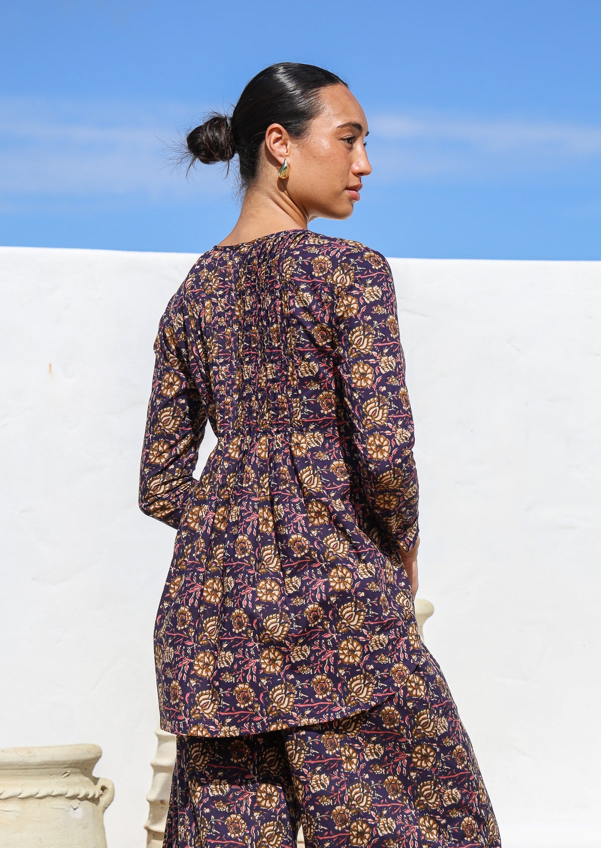 Woman wears cotton long sleeve top with small pin tucks across front and back of bodice, that is longer at the back.