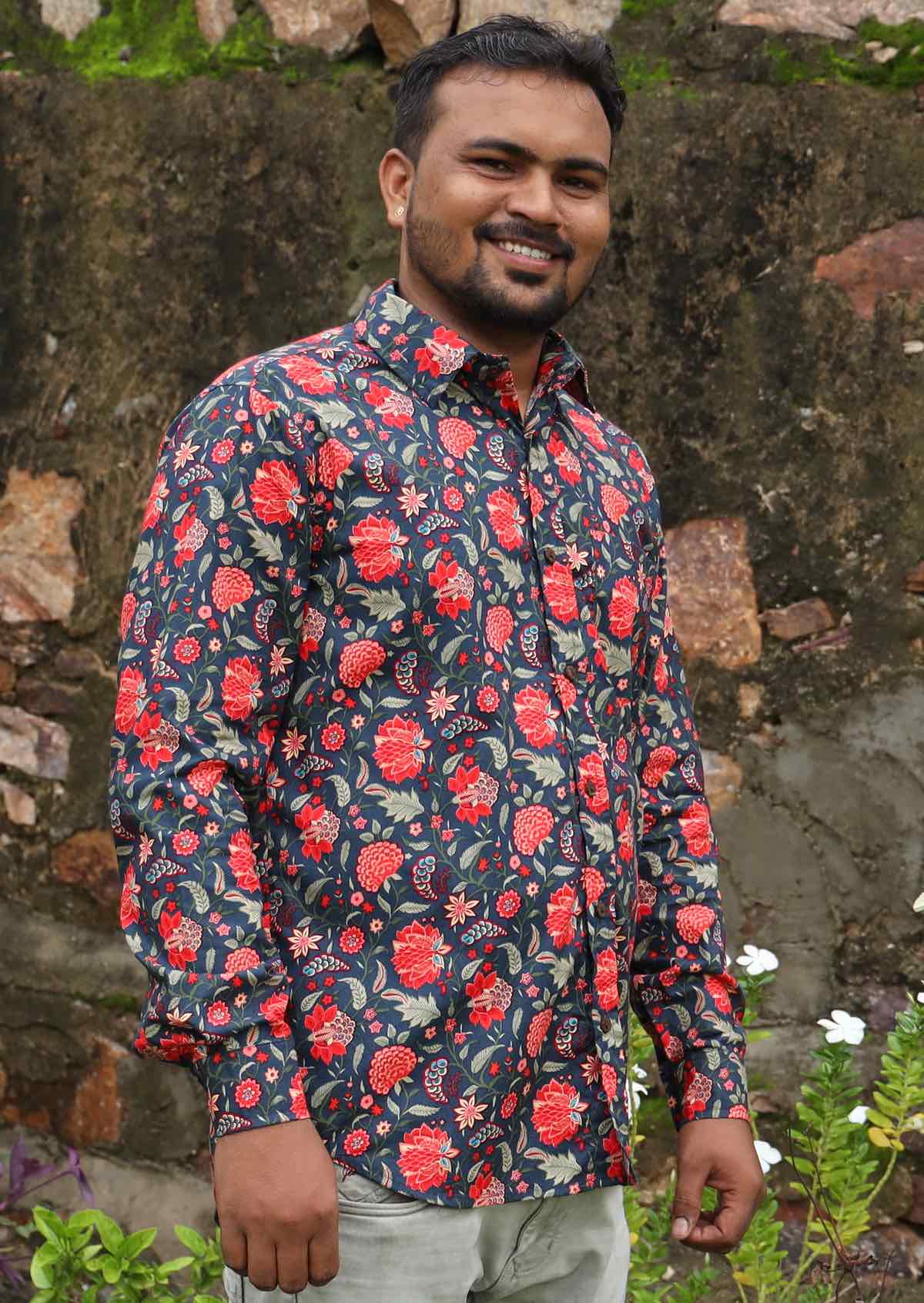Man wearing a floral print long sleeve shirt standing against a stone wall.