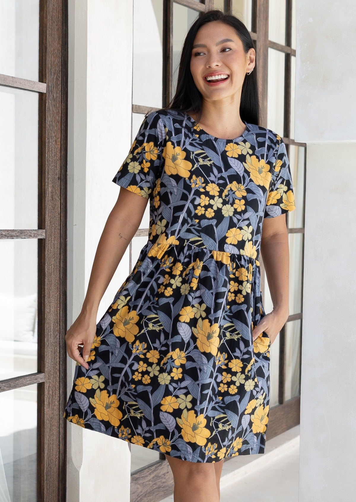 Woman wearing a black based blue-grey and yellow floral cotton dress, holding skirt out slightly