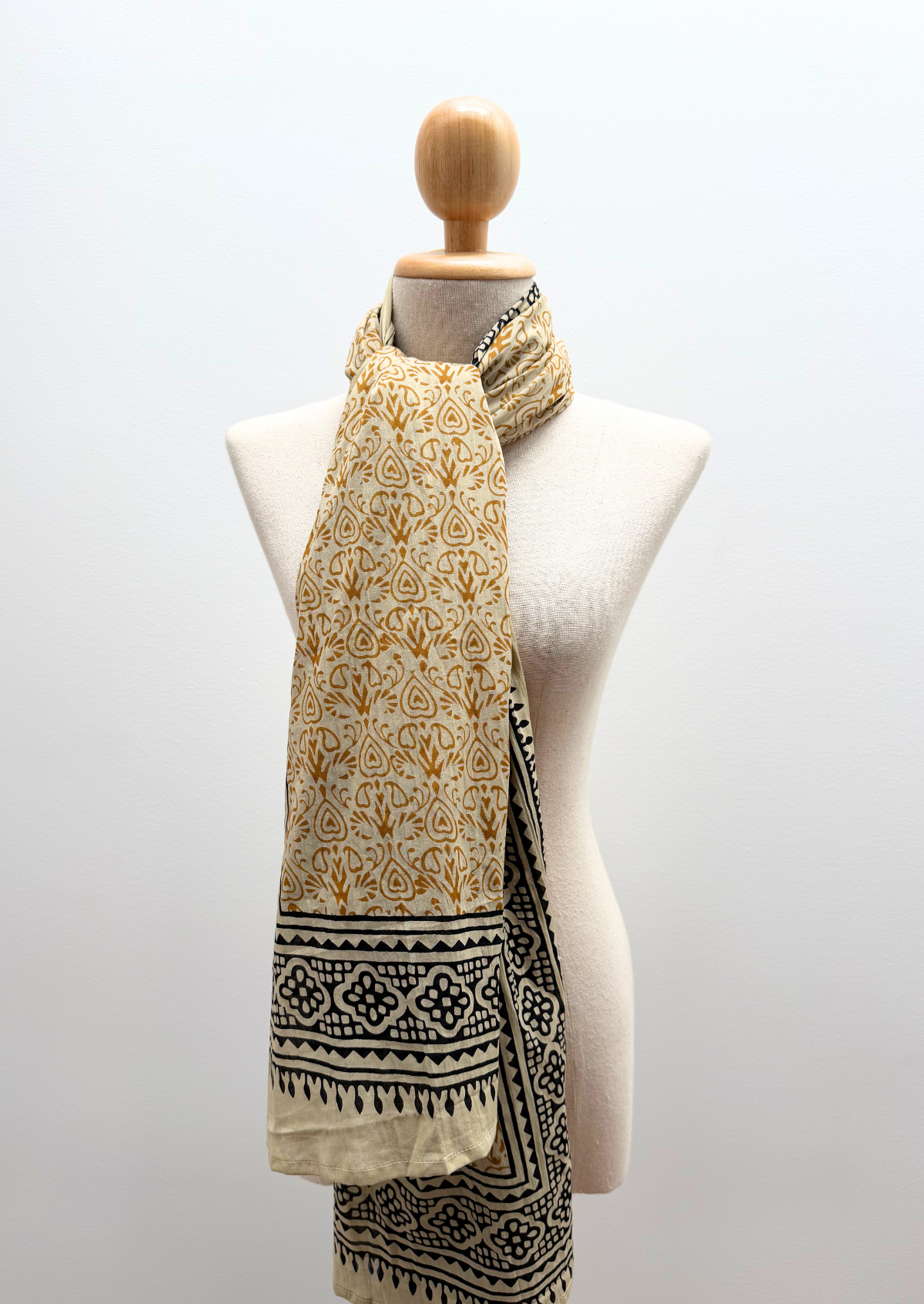 A lightweight scarf with a mustard yellow floral pattern and a black geometric border, draped on a cream-colored fabric mannequin torso against a light gray background.