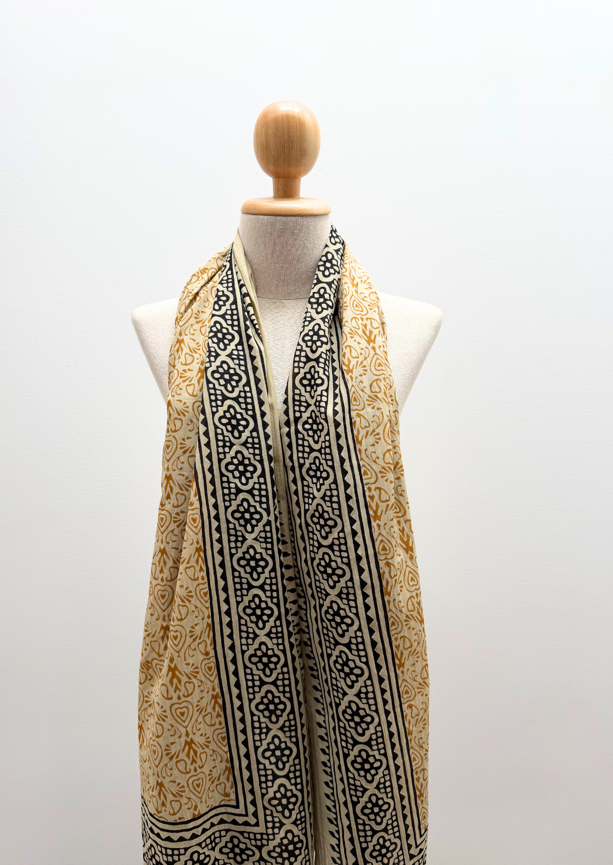 A lightweight scarf with a black and gold block-printed geometric pattern, draped on a cream-colored mannequin torso against a plain white background.