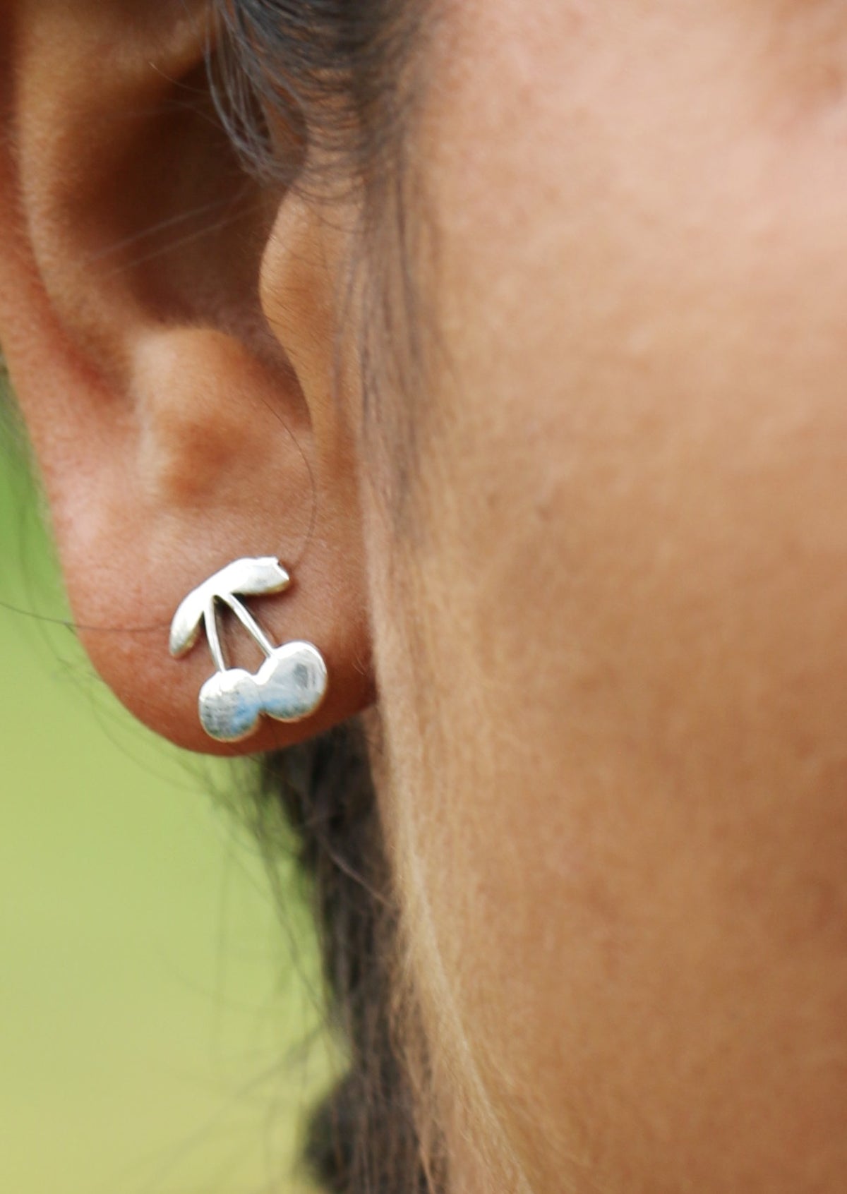 Close-up of an ear wearing a sterling silver cherry shaped studs with a blurred green background