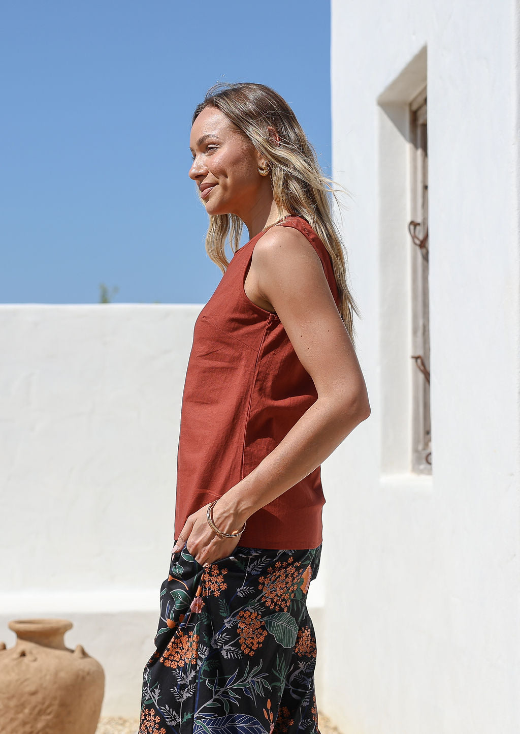 Woman wearing a relaxed fit rust-colored sleeveless cotton top with side bust darts and floral pants standing outdoors in front of white wall