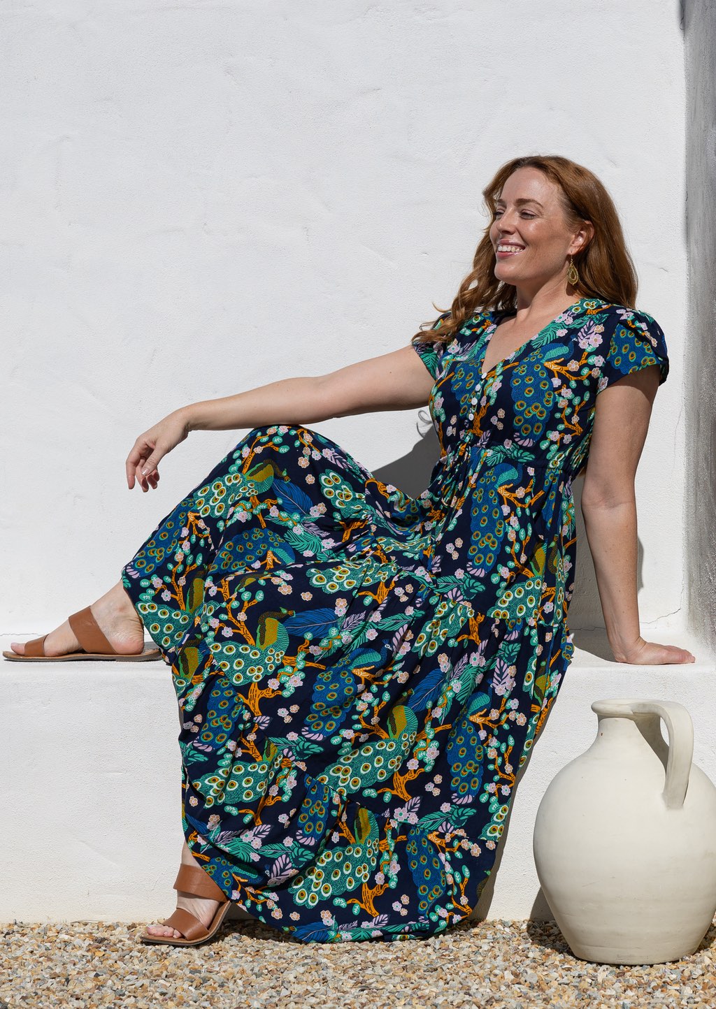 Woman wearing a cap-sleeve maxi dress with a colourful peacock print in teal, blue, yellow, and orange, sitting on a white stone ledge next to a white ceramic jug.