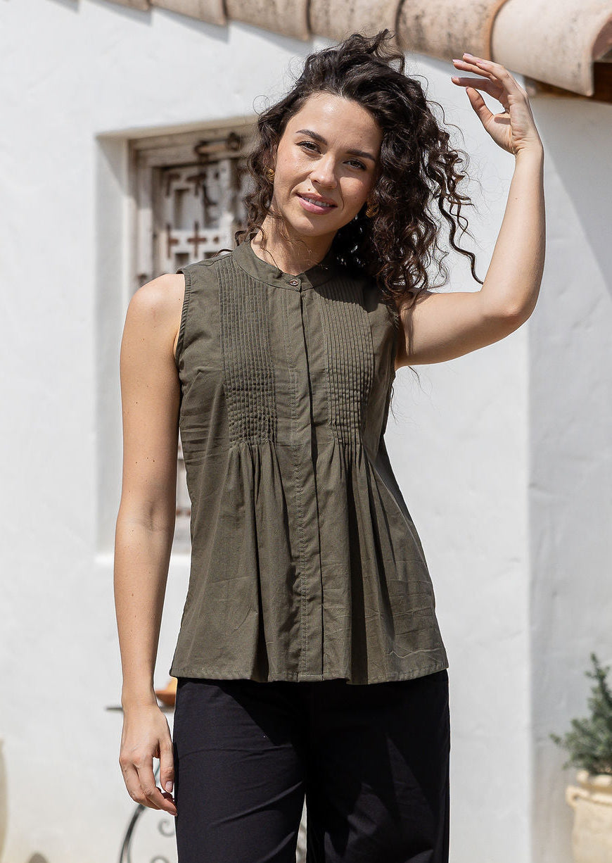 Woman wearing a lightweight 100% cotton olive green shade sleeveless top with pin tuck detailing and mandarin collar, standing in front of a rustic wooden window.