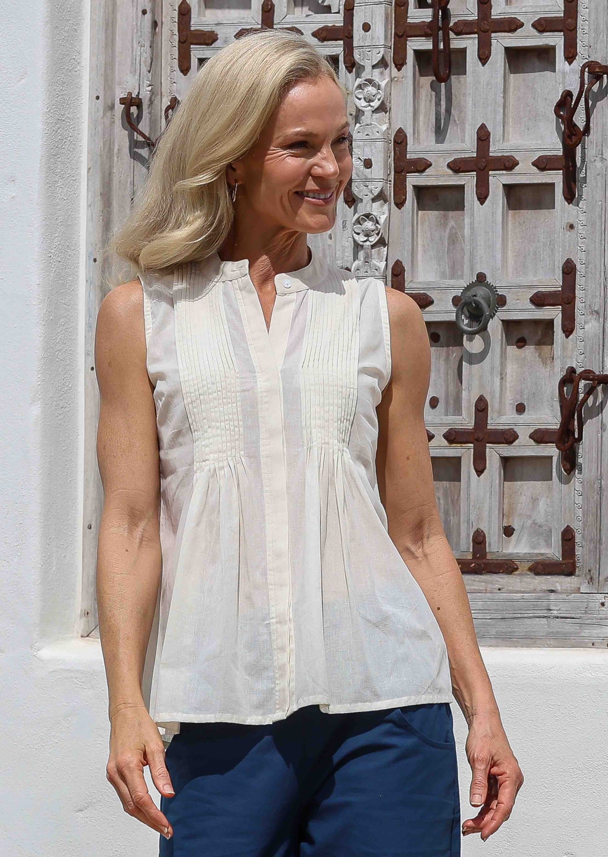 Woman wearing a white sleeveless cotton blouse with tiny pleats in front and blue pants standing in front of a decorative wooden window.