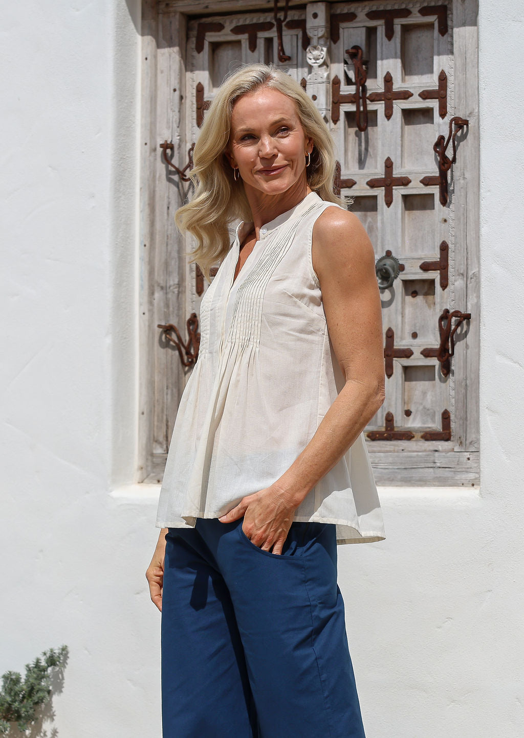 Woman wears 100% cotton natural white, sleeveless top with buttoned mandarin collar, round neckline and decorative small pleated design in front.