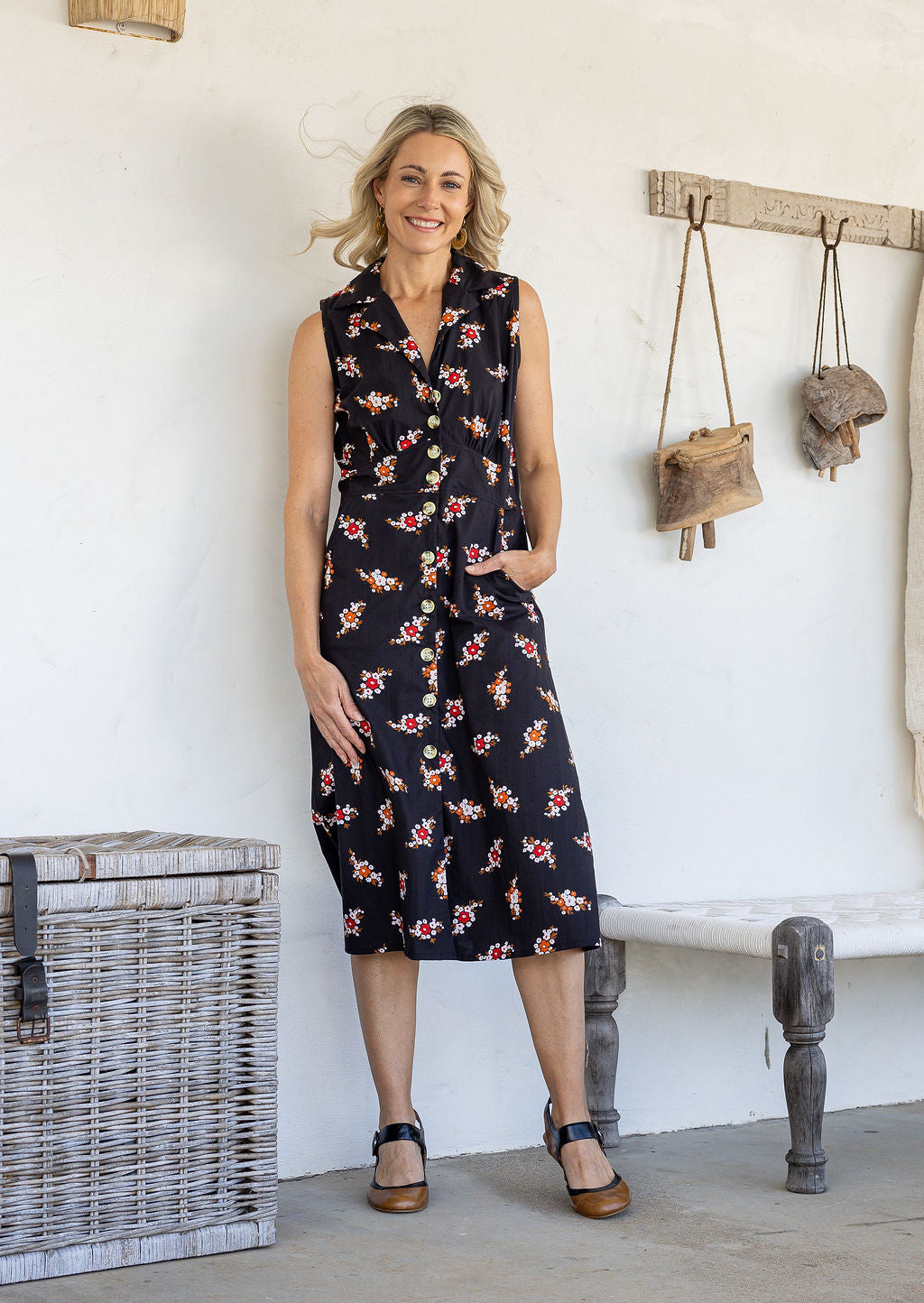 Woman wearing midi length, sleeveless cotton dress with collar and pockets in black based floral print.