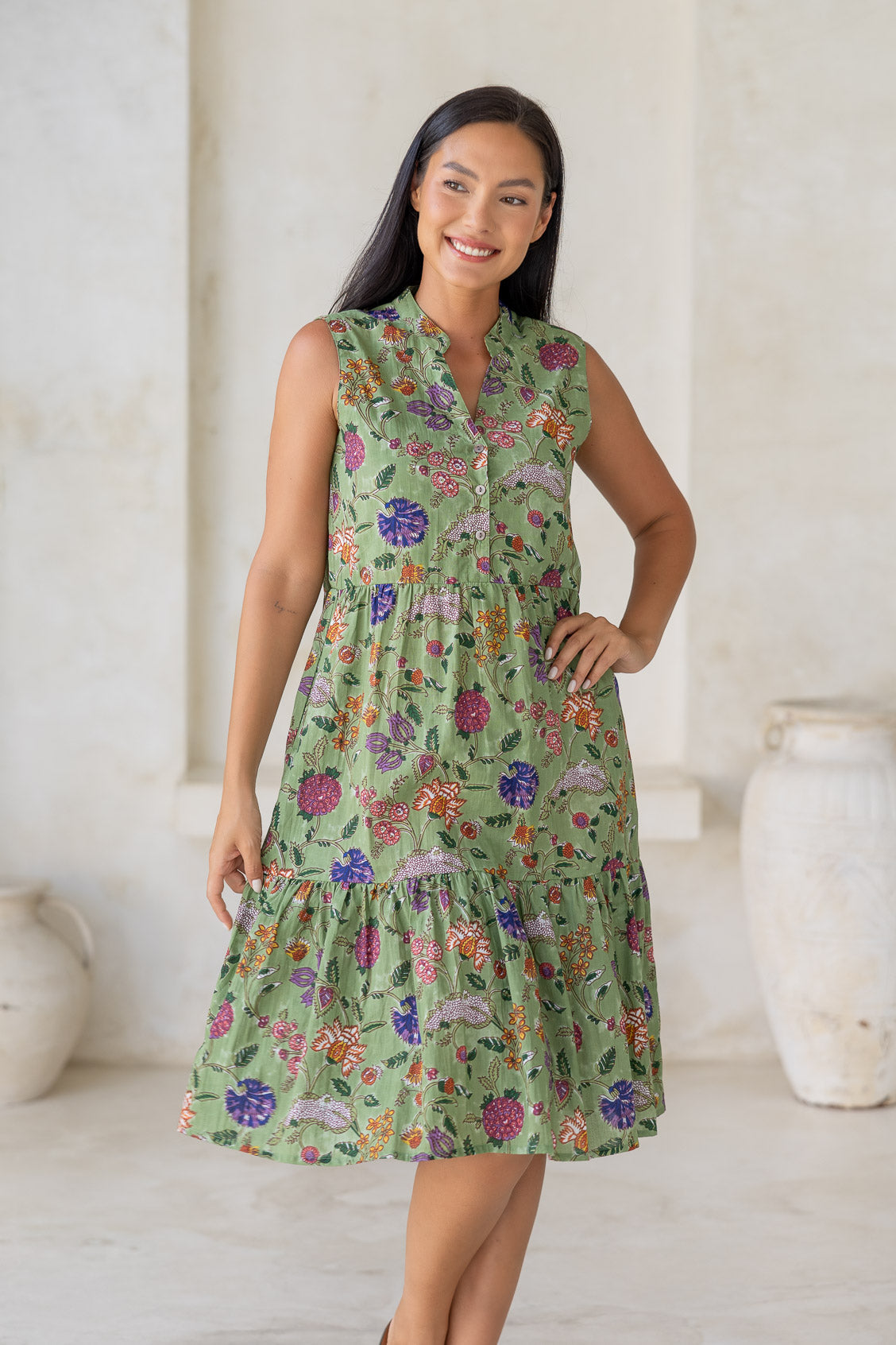 Woman wearing a green floral cotton dress with buttoned bodice and two tiered skirt, standing in a minimal indoor setting