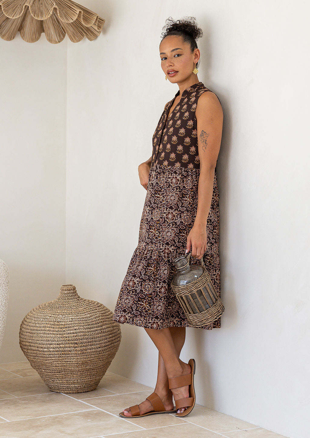 Woman in a brown 100% cotton patterned dress holding a woven basket against a white wall.