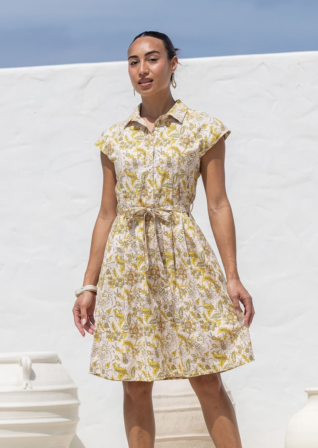 Woman wearing a 100% cotton yellow floral print shirt dress with collar, cap sleeves, and tie waist.