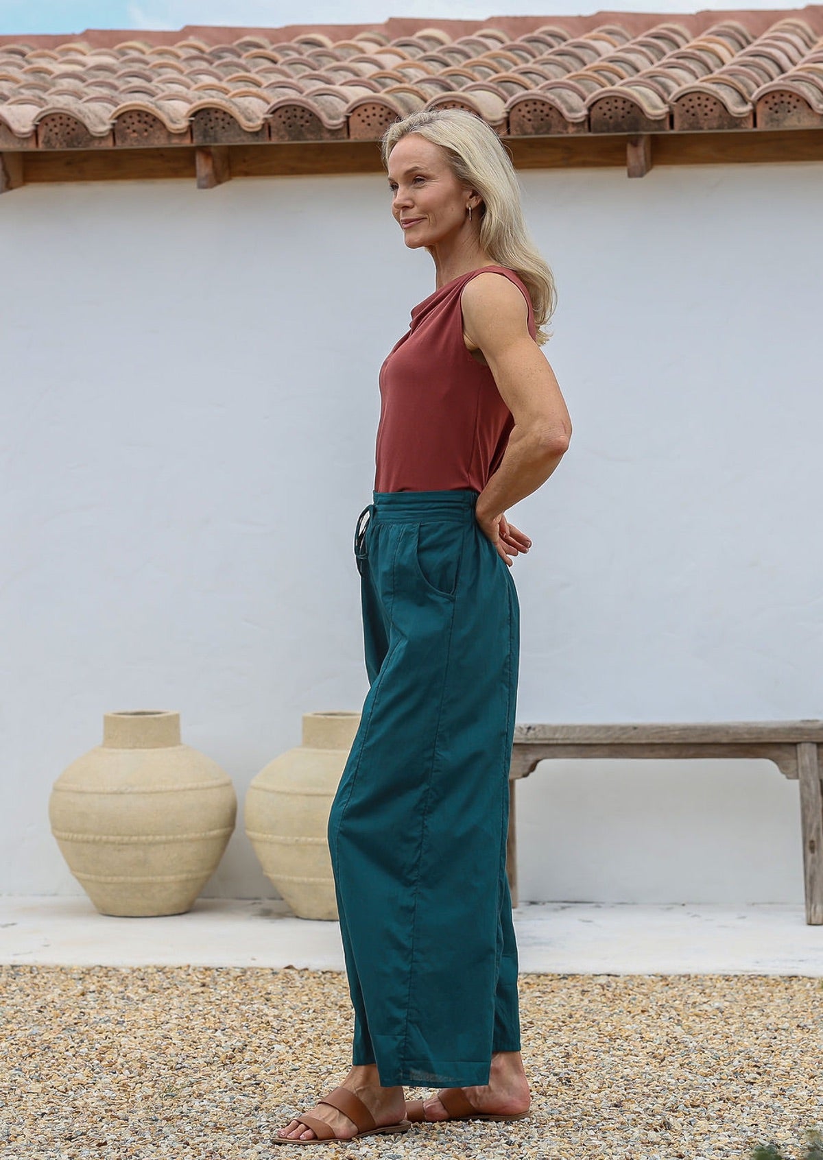 Woman wearing a rust top and deep teal 100% cotton, wide-legged pants standing outdoors with a white wall and wooden bench in the background.