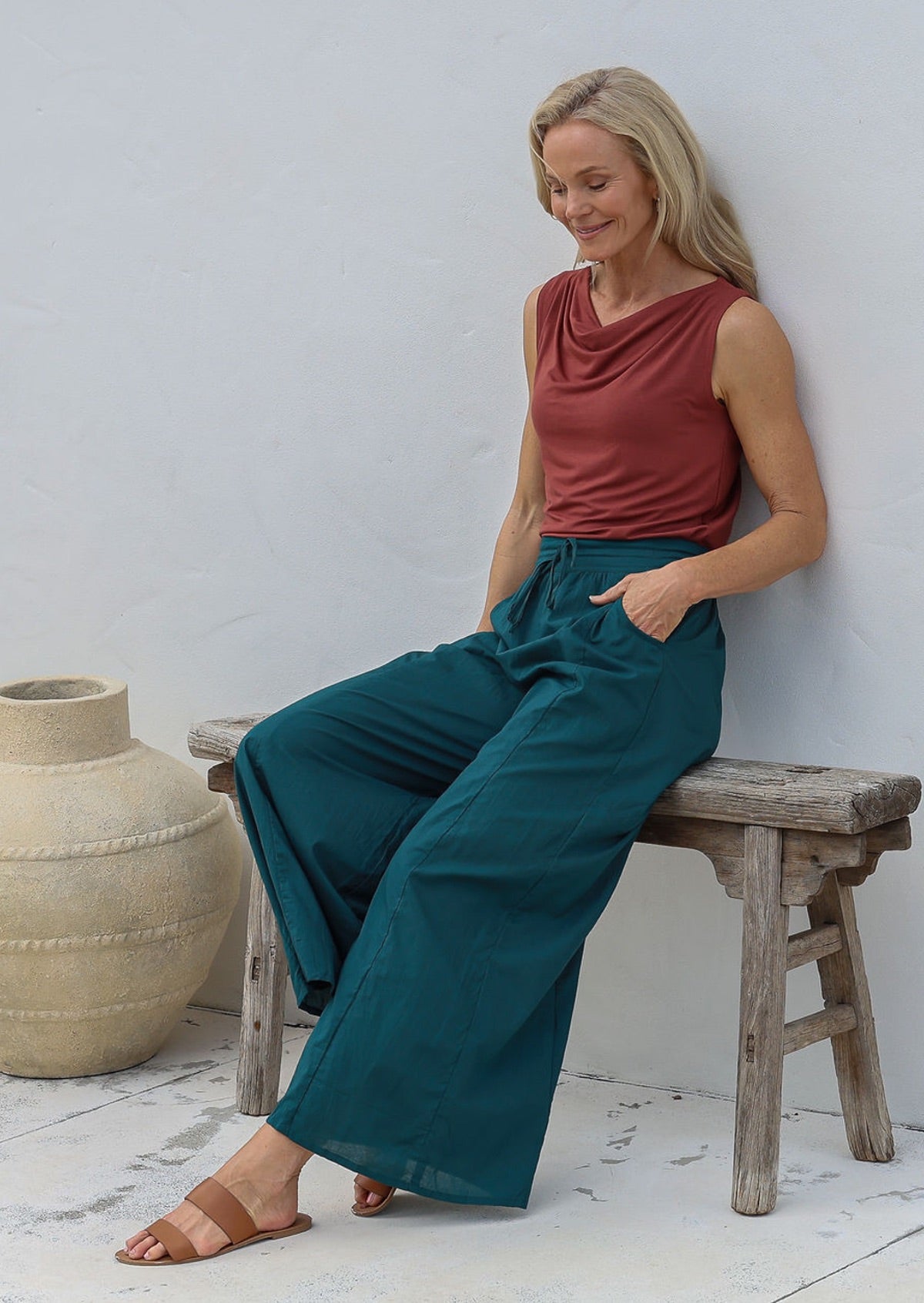 Woman sitting on a wooden stool wears 100% cotton wide leg pants in deep teal with pockets, elastic waist and drawstring tie, paired with terracotta basic top.