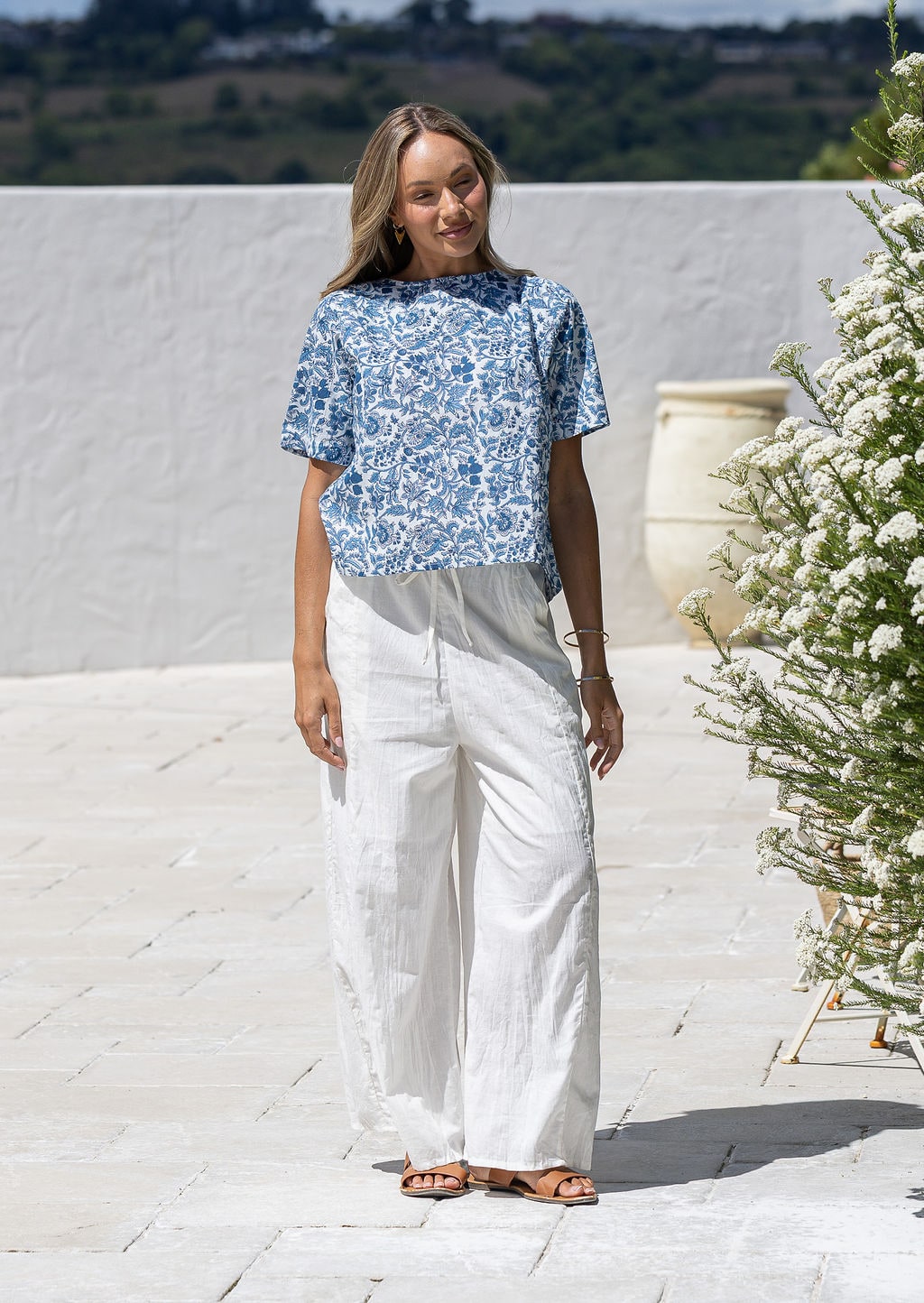 Woman wearing a white based blue floral printed cotton short sleeved top and white plain pants.