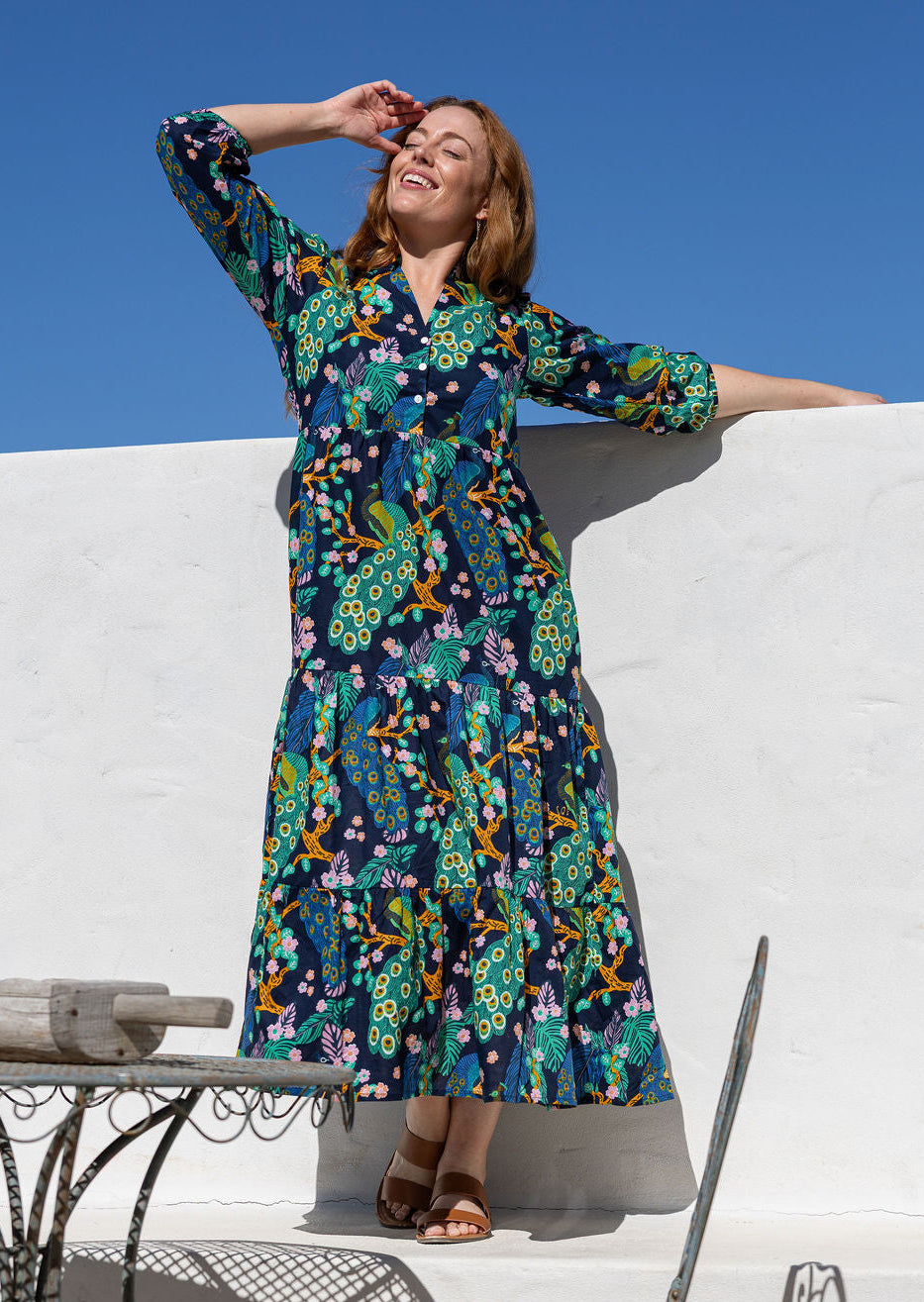 Woman wearing 3/4 sleeved maxi dress with a colourful floral and peacock print in shades of green, blue, yellow, and pink, posing outdoors against a white wall and blue sky.
