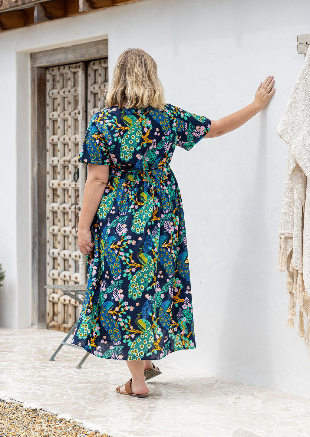 Woman standing backwards wearing a short-sleeve cotton midi length dress with a floral and peacock print on a dark navy background.