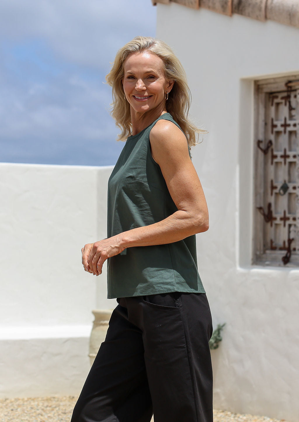 Woman wearing a jungle green sleeveless cotton top and black pants standing in front of a white wall with a window.