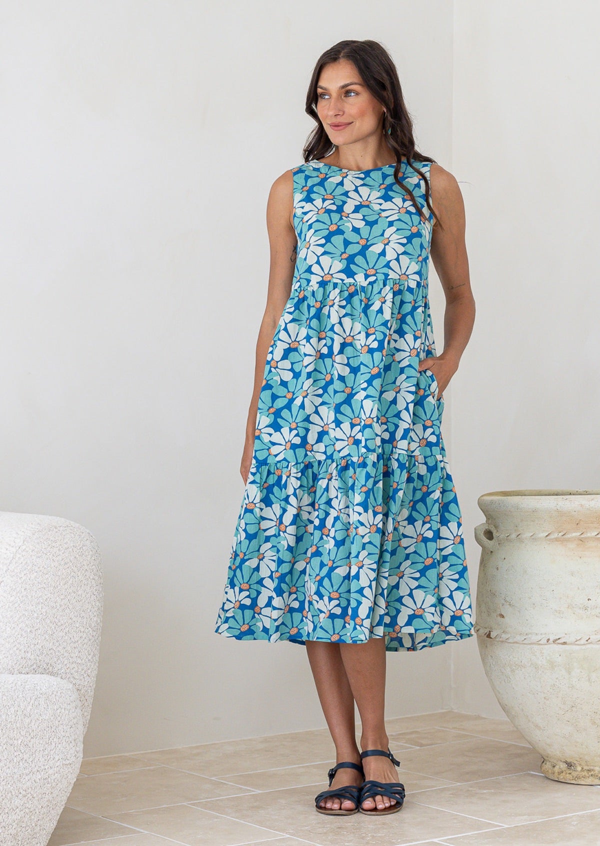 Woman poses in cotton sleeveless three tiered midi length dress in aqua and royal blue daisy print, with hand in pocket
