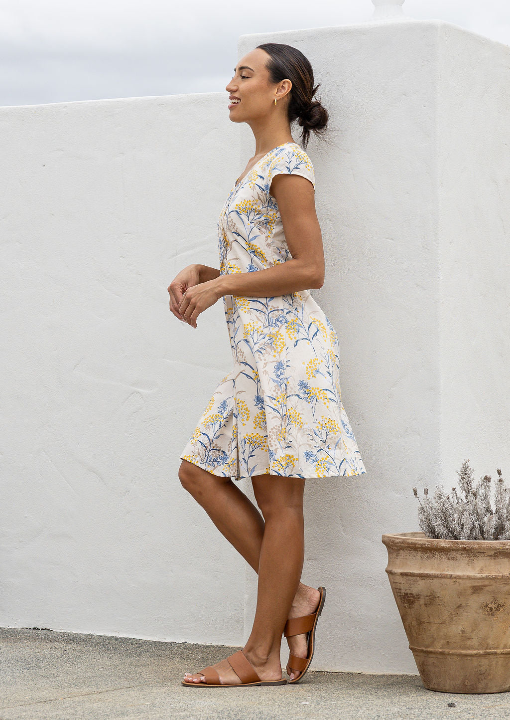 Woman in a floral dress standing against a white wall with a plant in the foreground.