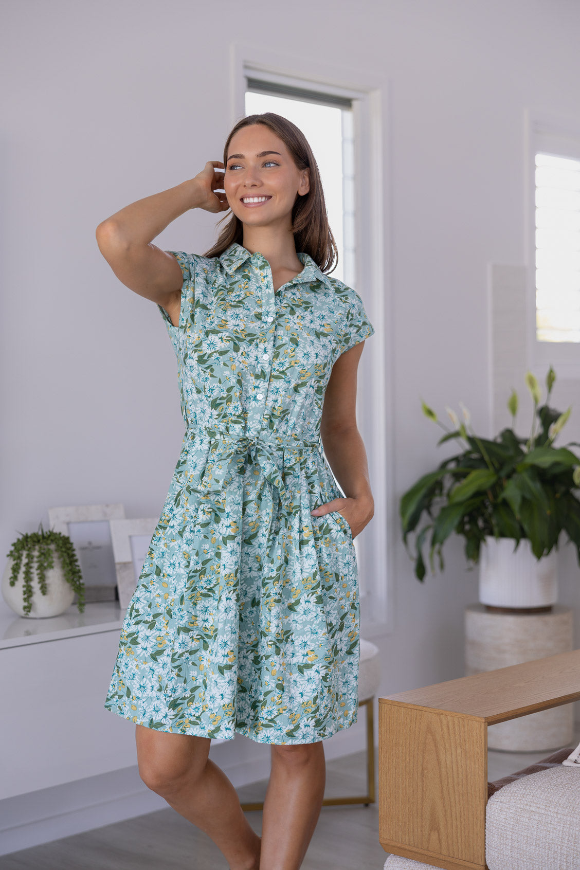 Woman wears fit and flare style shirt dress, 100% cotton with collar, cap sleeves, and removable waist tie in sage green based floral print.