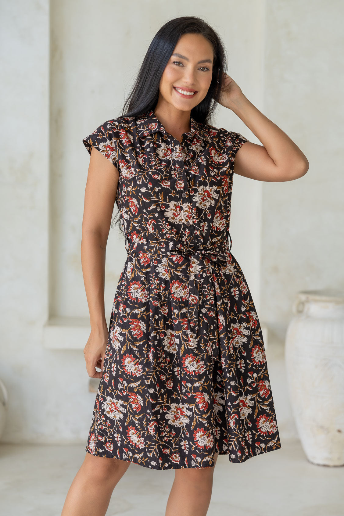 Woman wears fit and flare style shirt dress, 100% cotton with collar, cap sleeves, and removable waist tie in black based indian floral print.