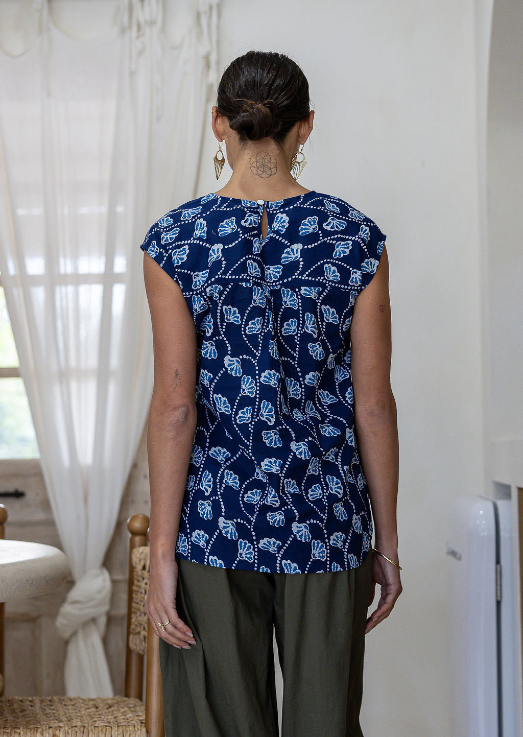 Woman stands backwards in a navy based floral sleeveless cotton top with button closure at the back paired with plain pants