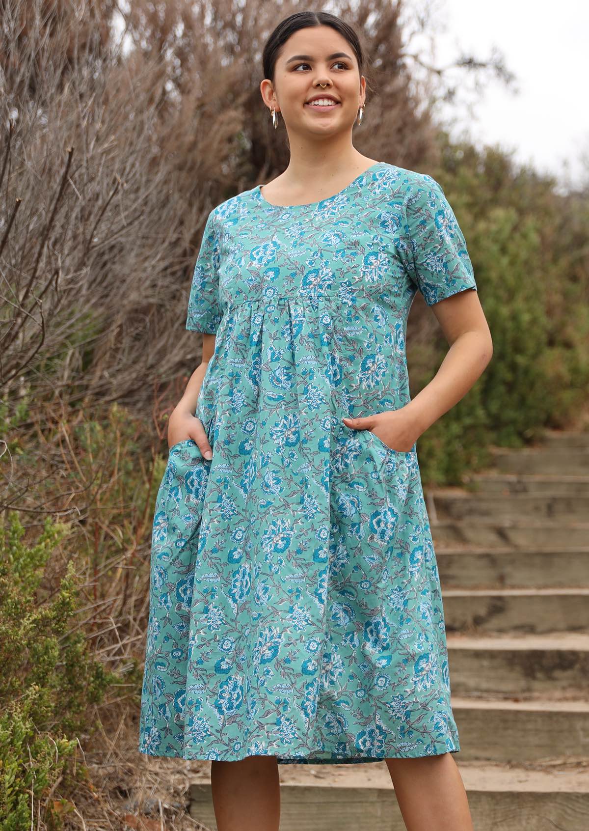 Model wears floral printed cotton dress in aqua shade with short sleeves and hands in pocket.