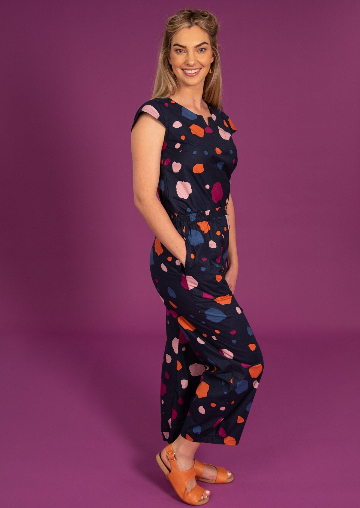 Woman stands in matching cotton top and pants in a multi coloured print on a navy blue base, giving a jumpsuit like appearance