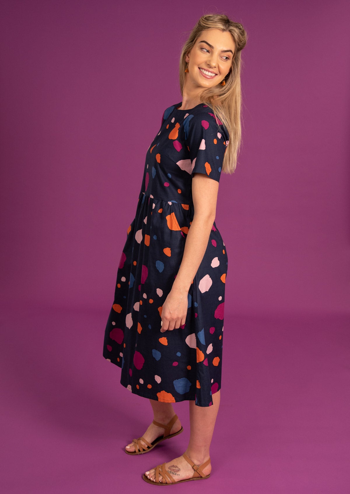 Model standing slightly sideways wears a beautiful bold spot print dress with short sleeves and pockets.