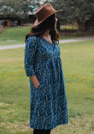 Woman wearing a blue floral dress and brown hat standing in a grassy area.