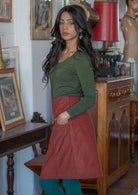 Woman stands sideways wearing a cotton corduroy A-line skirt with side zip with her hands in the pockets, and a long sleeve olive top