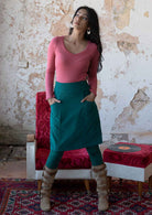 Woman wears cotton corduroy A-line knee length skirt in teal with hands in pockets, paired with a pink long sleeve top, leggings and boots