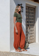 A woman wearing a green basic top and brick orange, genie style pull-on cotton pants with pockets, cuffed buttoned ankles, and a shirred elastic waistband.