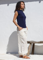 Woman wearing a navy basic top and 100% cotton white pull-on style pants with cuffed ankles and pockets, standing against a white wall with a wooden bench.