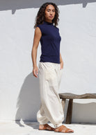 Woman wearing a navy sleeveless top and relaxed fit white cotton pants with pockets and buttoned cuffed ankles standing against a white wall.