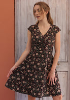 Woman stands in cotton above knee dress with cap sleeves and pockets, in bold maroon floral pattern on black base.
