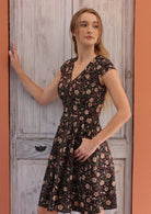 Woman stands sideways in retro style cotton above knee dress with cap sleeves and pockets, in floral print on black base.