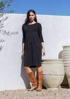 Woman wearing a black 100 cotton dress and brown boots standing against a white wall with decorative urns.