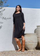 Woman in a black cotton dress and brown boots standing against a white wall with a blue sky.