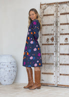 Woman stands side on in a navy blue base dress with colourful polka dots and tan knee-high boots