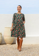 Woman wears 100% cotton knee length dress in deep green with beautiful pinkish red floral print and hand in pocket.