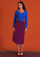 woman wearing purple corduroy aline skirt  with belt loops with bright blue long sleeve top standing on persian carpet

