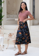 Woman standing side on in a black based cotton floral midi length skirt, paired with a  soft pink rayon top