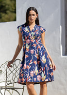 Woman standing against the wall wears floral print navy blue dress with A line skirt and box pleats from the waist.